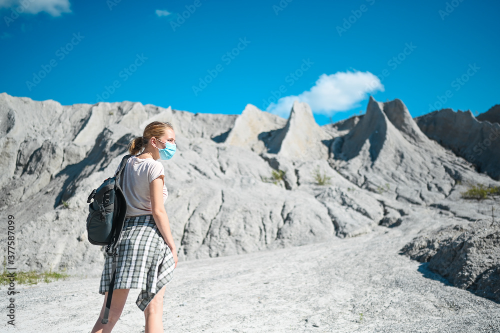 Young blonde woman walking against the white mountains wearing face protective mask for Covid 19 prevention. Young student traveler tourist with backpack. Corona virus and domestic tourism concept
