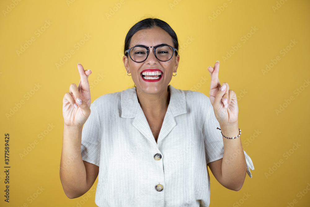 Beautiful woman over yellow background gesturing finger crossed smiling with hope and looking side Beautiful woman over yellow background gesturing finger crossed smiling with hope and looking side
