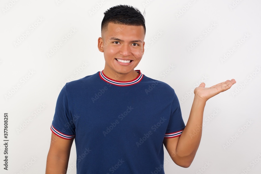 Young handsome hispanic man wearing casual t-shirt standing over white isolated background smiling cheerful presenting and pointing with palm of hand looking at the camera. Young handsome hispanic man wearing casual t-shirt standing over white isolated background smiling cheerful presenting and pointing with palm of hand looking at the camera.