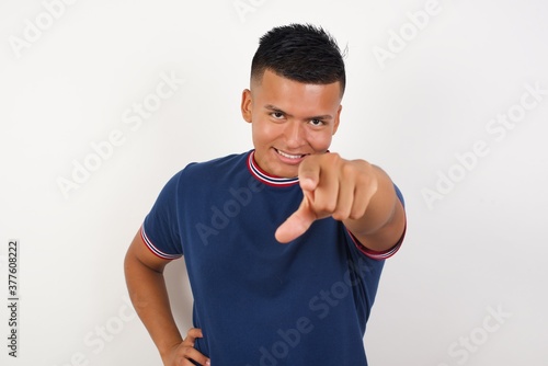 Young handsome hispanic man wearing casual t-shirt standing over white isolated background pointing at camera with a satisfied, confident, friendly smile, choosing you