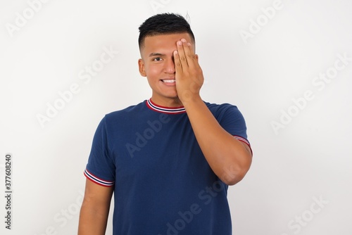 Young handsome hispanic man wearing casual t-shirt standing over white isolated background covering one eye with her hand, confident smile on face and surprise emotion.