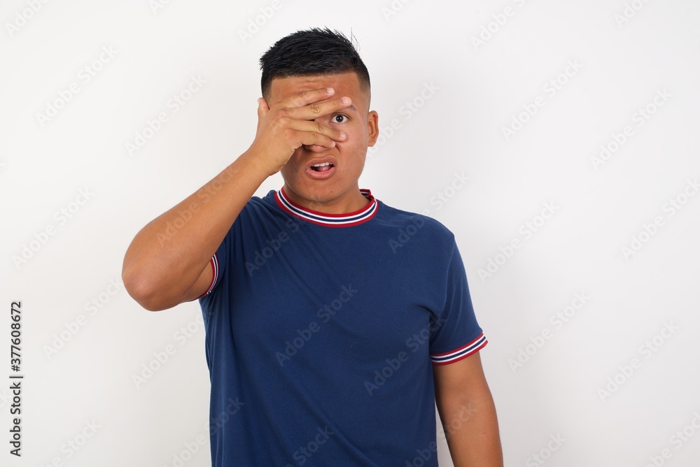 Young handsome hispanic man wearing casual t-shirt standing over white isolated background peeking in shock covering face and eyes with hand, looking through fingers with embarrassed expression. Young handsome hispanic man wearing casual t-shirt standing over white isolated background peeking in shock covering face and eyes with hand, looking through fingers with embarrassed expression.