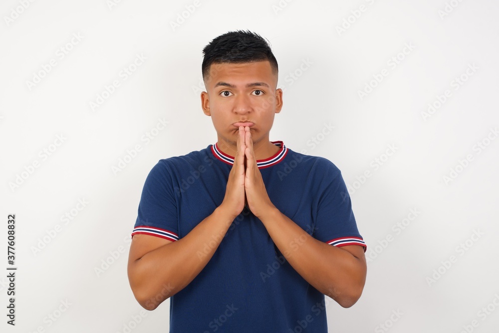 Positive Young handsome hispanic man wearing casual t-shirt standing over white smiles happily, glad to receive pleasant news from interlocutor, keeps palms together, People emotions concept. Positive Young handsome hispanic man wearing casual t-shirt standing over white smiles happily, glad to receive pleasant news from interlocutor, keeps palms together, People emotions concept.