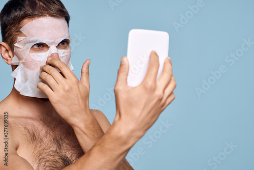 a man with a cleansing mask on his face examines himself in a mirror on a blue background
