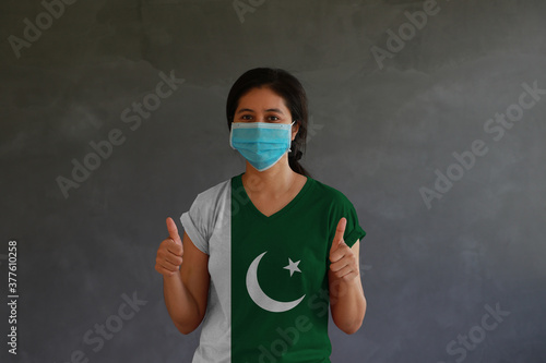 Masked woman wearing Pakistan flag color of shirt and thumbs up with both hands on dark wall background. photo