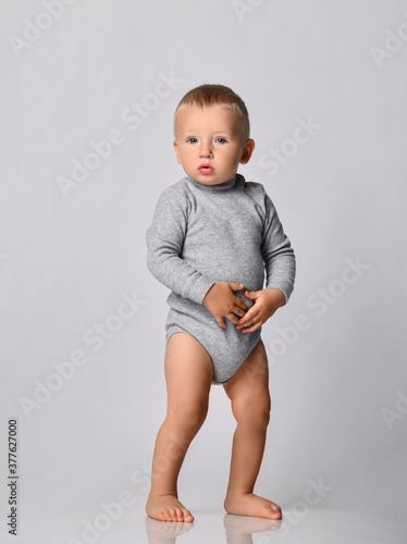 Toddler one-year-old baby boy in diaper and grey one-piece bodysuit with long sleeves stands looking at camera