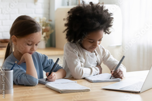 Focused small diverse sisters sitting at table, doing tasks in notebooks together at home. Busy little mixed race kids girls involved in preparing school homework or enjoying online educational class.