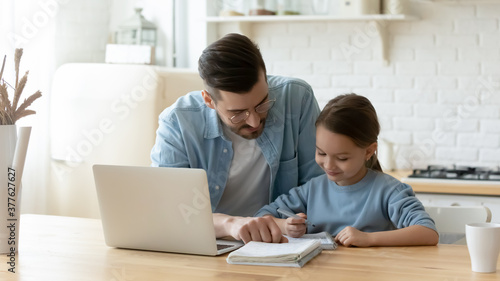 Caring young dad helping small primary pupil kid daughter preparing school homework, sitting together at table. Happy little child girl involved in doing task, studying homeschooling with father.