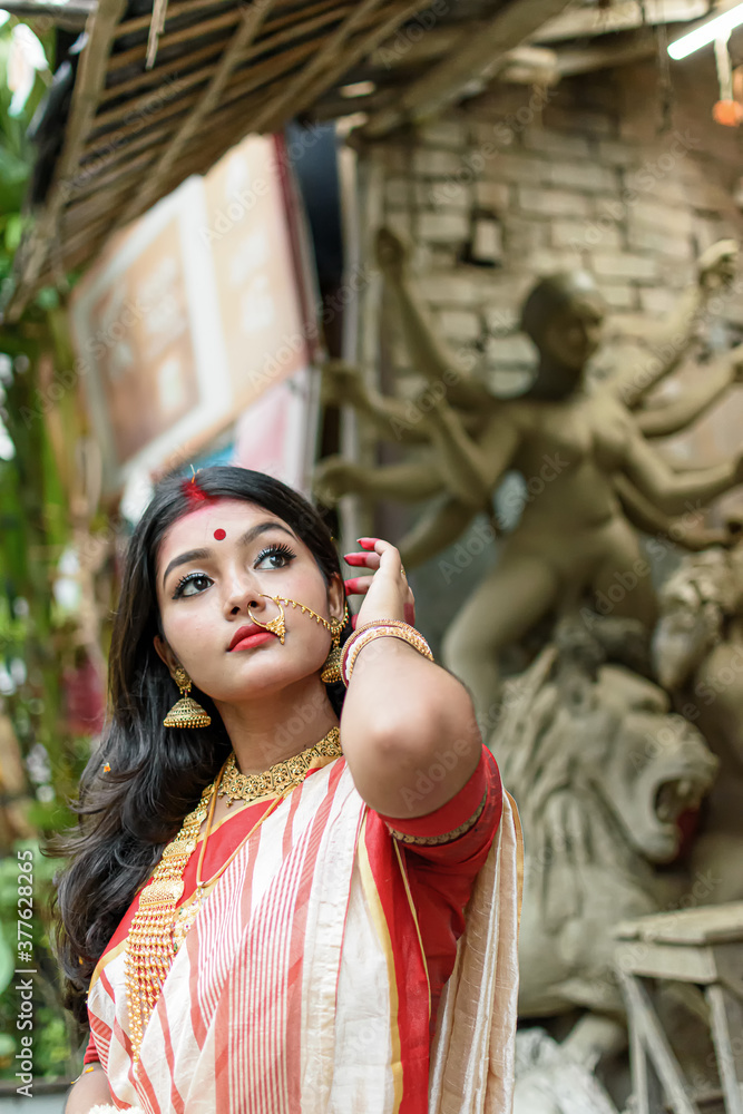 Portrait of stunning Indian woman draped in traditional saree adorned with gold jewellery, posing before the clay idol of the goddess Devi Durga Portrait of stunning Indian woman draped in traditional saree adorned with gold jewellery, posing before the clay idol of the goddess Devi Durga