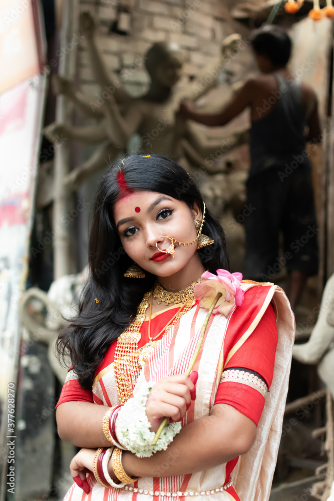 Obraz premium Portrait of stunning Indian woman draped in traditional saree adorned with gold jewellery, posing before the clay idol of the goddess Devi Durga