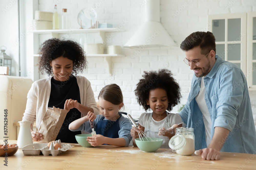 Happy multiracial young couple involved in cooking pastry with adorable mixed race daughters in modern kitchen. Smiling diverse family preparing homemade sweets or pancakes for breakfast at weekend. Happy multiracial young couple involved in cooking pastry with adorable mixed race daughters in modern kitchen. Smiling diverse family preparing homemade sweets or pancakes for breakfast at weekend.