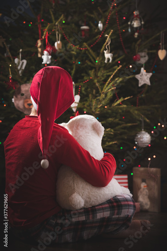 A cute toddler boy sits next to an evergreen fir tree decorated with garlands and other New Year's toys, waiting for the holiday on a winter evening. Holiday and magic concept