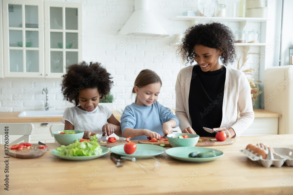 Affectionate caring african american mommy teaching little preschool adorable mixed race kids girls chopping fresh vegetables for healthy salad food, standing together at countertop in modern kitchen. Affectionate caring african american mommy teaching little preschool adorable mixed race kids girls chopping fresh vegetables for healthy salad food, standing together at countertop in modern kitchen.