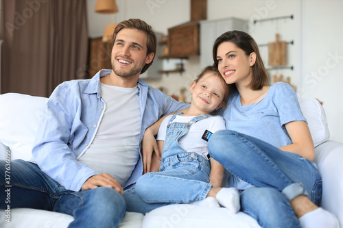 Happy family with child sitting on sofa watching tv, young parents embracing daughter relaxing on couch together.