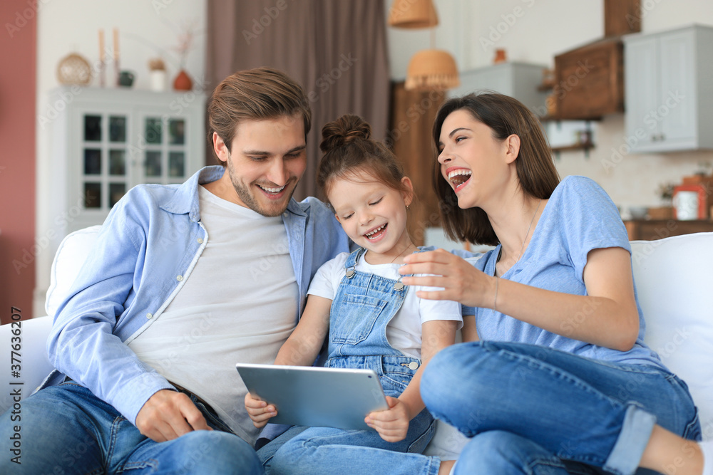 Positive friendly young parents with smiling little daughter sitting on sofa together answering video call on digital tablet while relaxing at home on weekend. Positive friendly young parents with smiling little daughter sitting on sofa together answering video call on digital tablet while relaxing at home on weekend.