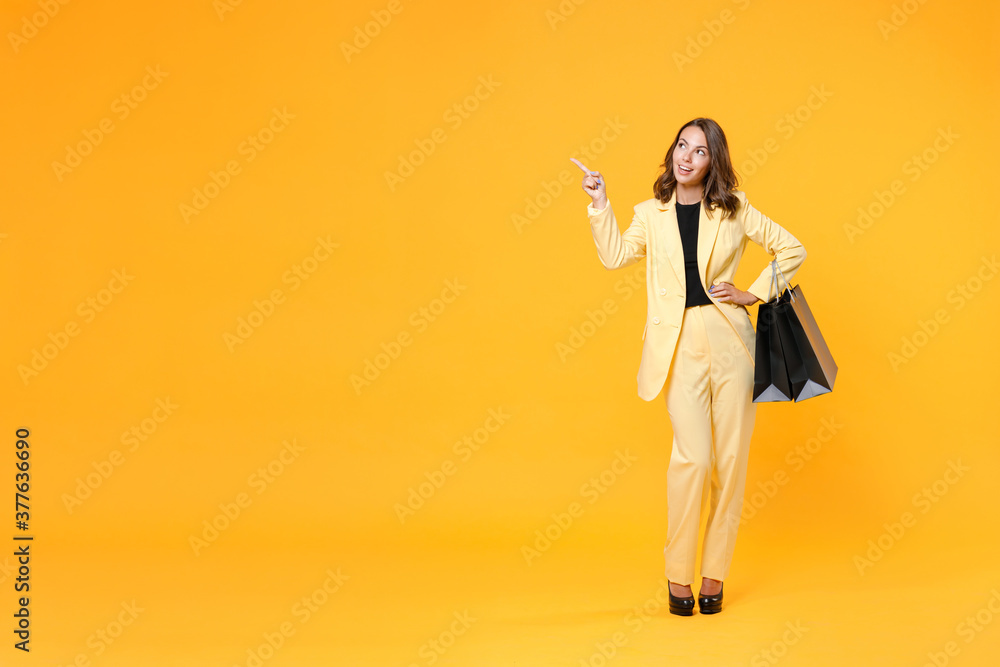 Full length portrait of smiling young woman in suit jacket hold package bags with purchases after shopping point finger aside up on mock up copy space isolated on yellow background. Black friday sale. Full length portrait of smiling young woman in suit jacket hold package bags with purchases after shopping point finger aside up on mock up copy space isolated on yellow background. Black friday sale.