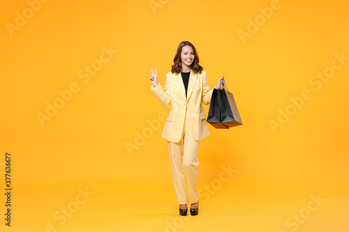 Full length portrait of smiling young woman 20s in basic light suit jacket hold package bags with purchases after shopping showing victory sign isolated on yellow background studio. Black friday sale.