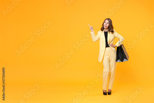 Full length portrait of smiling young woman in suit jacket hold package bags with purchases after shopping point finger aside up on mock up copy space isolated on yellow background. Black friday sale. © ViDi Studio