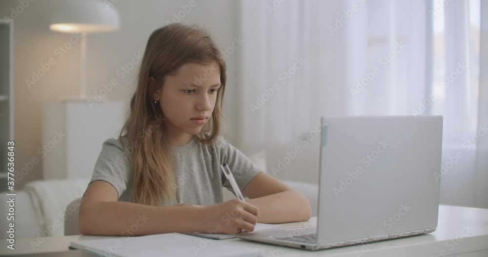 teen girl is learning distantly, writing in exercise book and looking at display of notebook, staying home teen girl is learning distantly, writing in exercise book and looking at display of notebook, staying home