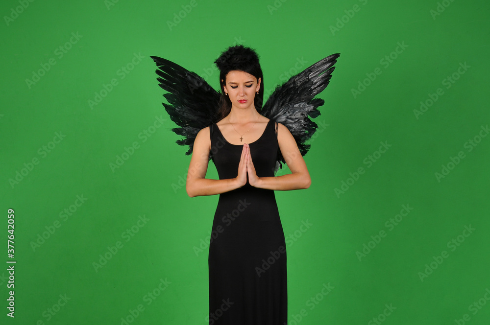 Beautiful young brunette woman in black angel costume with wings over green background in praying pose. Halloween party outfit Beautiful young brunette woman in black angel costume with wings over green background in praying pose. Halloween party outfit