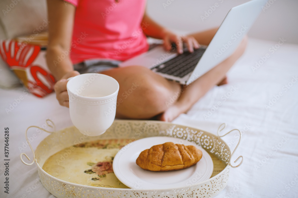 Caucasian girl  sitting on the bed working on a laptop, having breakfast in bed. Work from home, stay home concept, home office, working home, self-isolation.