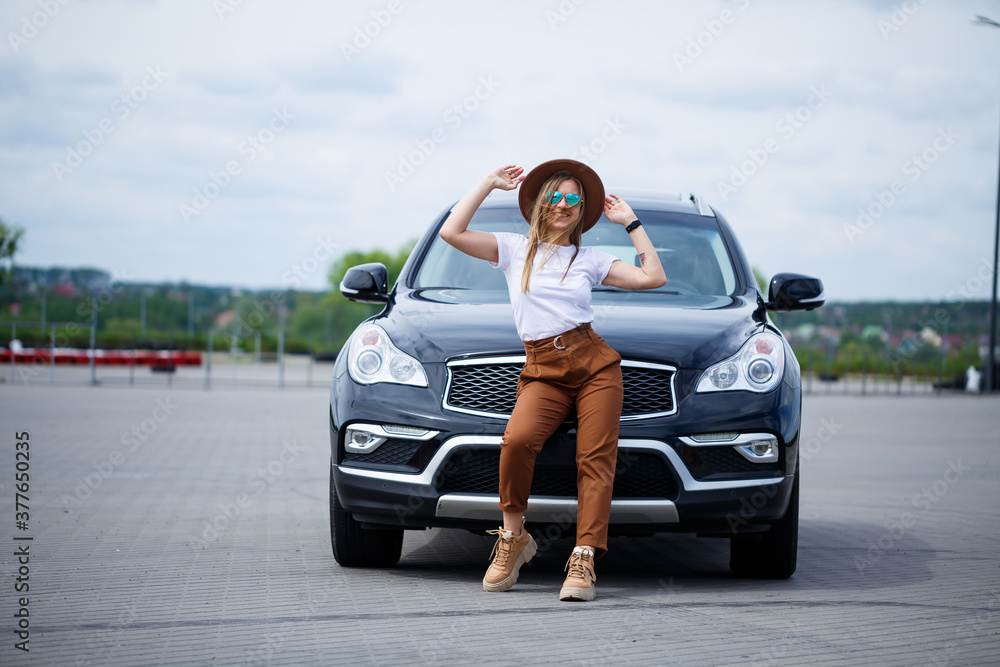 A beautiful girl of European appearance with glasses and a brown hat is standing near a black car. Young woman with car in parking A beautiful girl of European appearance with glasses and a brown hat is standing near a black car. Young woman with car in parking