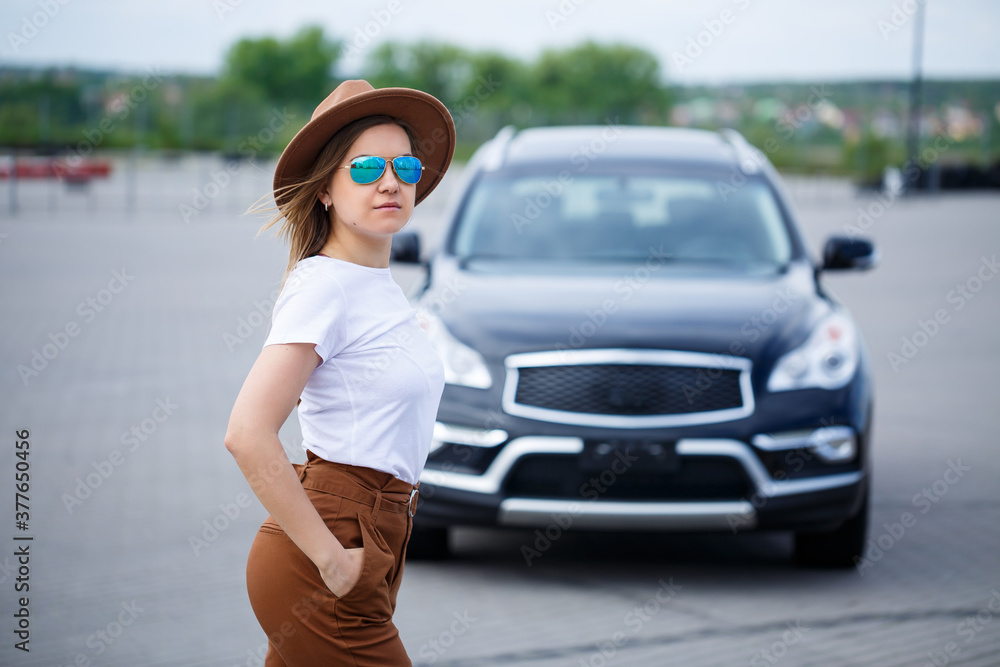Fototapeta premium A beautiful girl of European appearance with glasses and a brown hat is standing near a black car. Young woman with car in parking