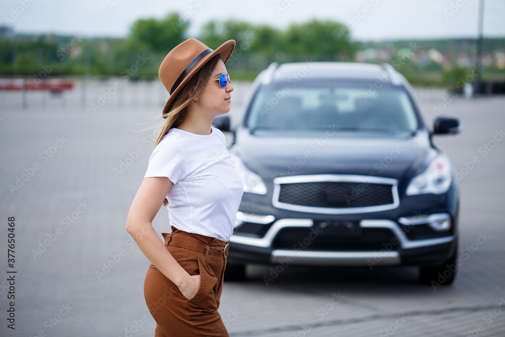 A beautiful girl of European appearance with glasses and a brown hat is standing near a black car. Young woman with car in parking