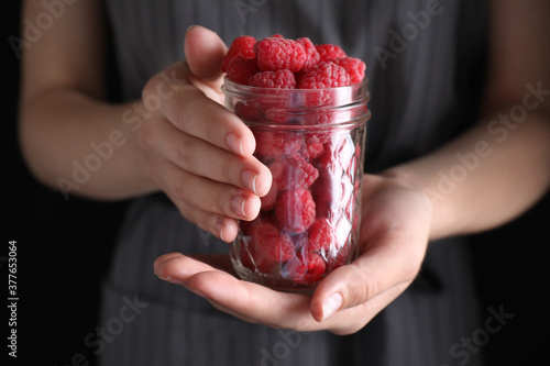 Woman holding glass jar of delicious ripe raspberries on black background, closeup