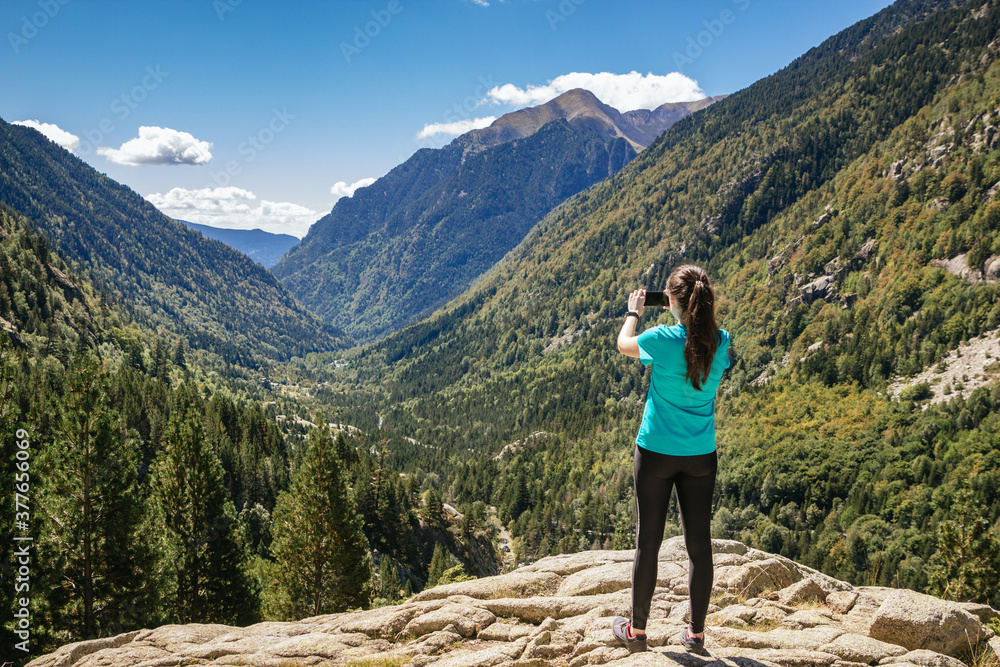 Stock photo of a woman with mask standing on a rock and taking a landscape photo with her phone