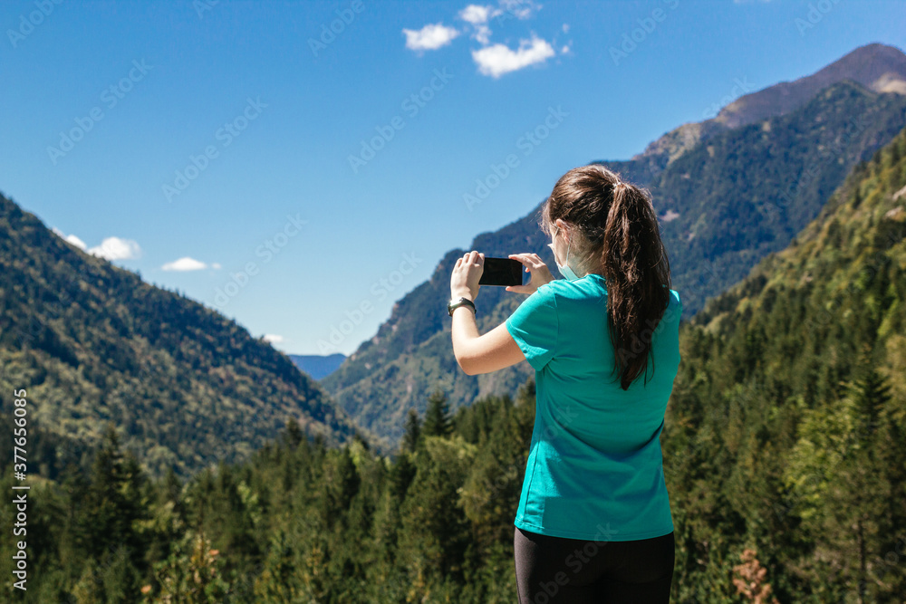 Stock photo of a woman with face mask taking a photo of a beautiful mountain landscape with her smartphone
