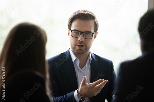 Serious Caucasian young businessman in glasses discuss business ideas with diverse colleagues at team meeting in office. Millennial male boss CEO talk with partners cooperate brainstorm at briefing.