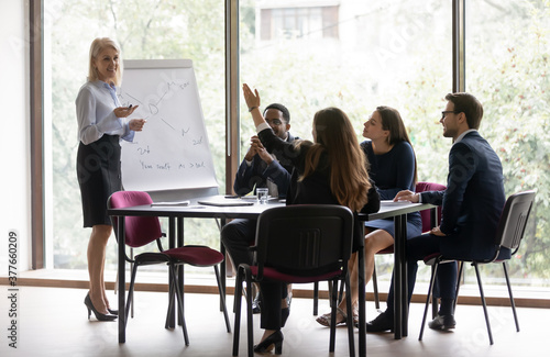 Middle-aged businesswoman make whiteboard presentation interact with diverse colleagues at meeting. Smiling mature female coach or trainer present project on flip chart  brainstorm with coworkers.