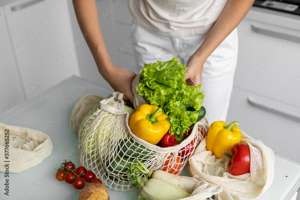Woman came back from a market and unpacks a reusable grocery bag full of vegetables on a kitchen at home. Zero waste and plastic free concept. Girl is holding mesh cotton shopper with vegetables.