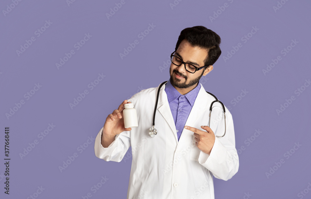 Confident Indian doctor pointing at jar of pills on lilac background, mockup for design Confident Indian doctor pointing at jar of pills on lilac background, mockup for design