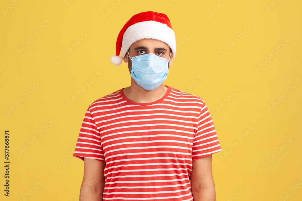 Serious man in red t-shirt and santa claus hat with surgical medical mask on face standing and looking at camera. Indoor studio shot isolated on yellow background Serious man in red t-shirt and santa claus hat with surgical medical mask on face standing and looking at camera. Indoor studio shot isolated on yellow background