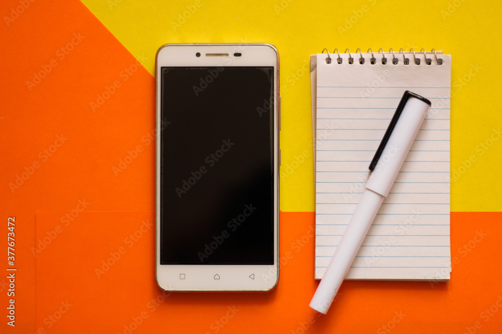 Creative flat lay, photo of workspace desk with smartphone, notepad, pen with a copy space, orange and yellow background, minimal style, selective focus Creative flat lay, photo of workspace desk with smartphone, notepad, pen with a copy space, orange and yellow background, minimal style, selective focus