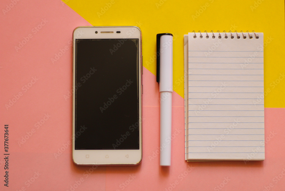 Creative flat lay, photo of workspace desk with smartphone, notepad, a pen with a copy space, pink and yellow background, minimal style, selective focus Creative flat lay, photo of workspace desk with smartphone, notepad, a pen with a copy space, pink and yellow background, minimal style, selective focus