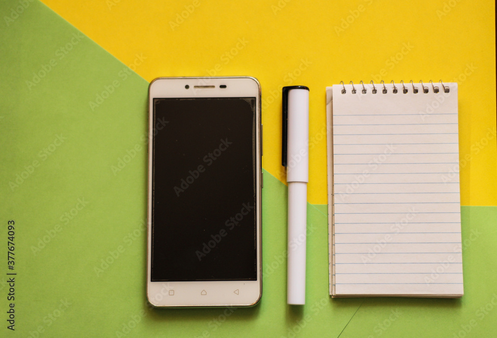 Creative flat lay, photo of workspace desk with smartphone, notepad, pen with a copy space, green and yellow background, minimal style, selective focus Creative flat lay, photo of workspace desk with smartphone, notepad, pen with a copy space, green and yellow background, minimal style, selective focus
