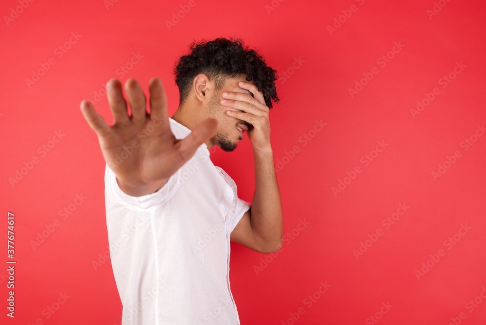 People, body language. Young arab man with afro hair covers eyes with palm and doing stop gesture, tries to hide from everybody. Don't look at me, I don't want to see, feels ashamed or scared. People, body language. Young arab man with afro hair covers eyes with palm and doing stop gesture, tries to hide from everybody. Don't look at me, I don't want to see, feels ashamed or scared.