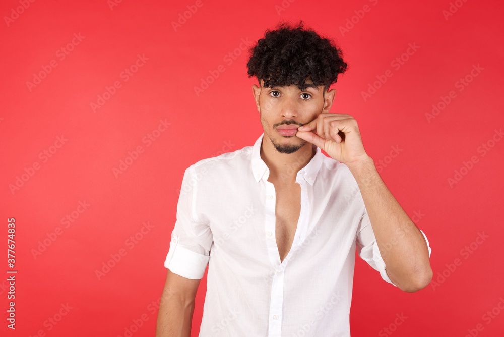 Young arab man with afro hair wearing shirt standing over isolated red background mouth and lips shut as zip with fingers. Secret and silent, taboo talking.