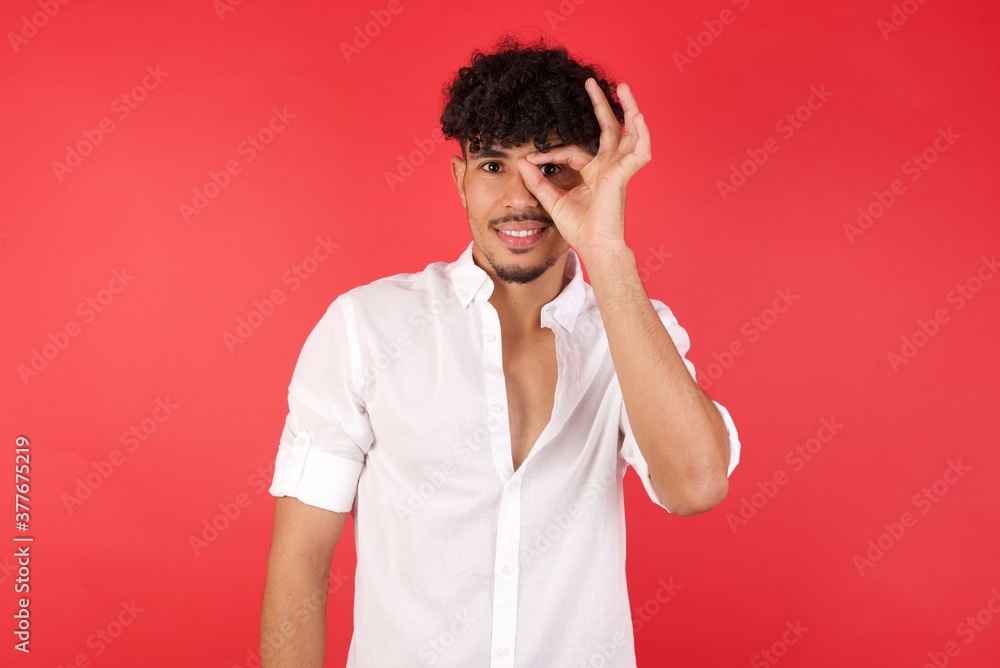 Young arab man with afro hair wearing shirt standing over isolated red background doing ok gesture with hand smiling, eye looking through fingers with happy face. Young arab man with afro hair wearing shirt standing over isolated red background doing ok gesture with hand smiling, eye looking through fingers with happy face.