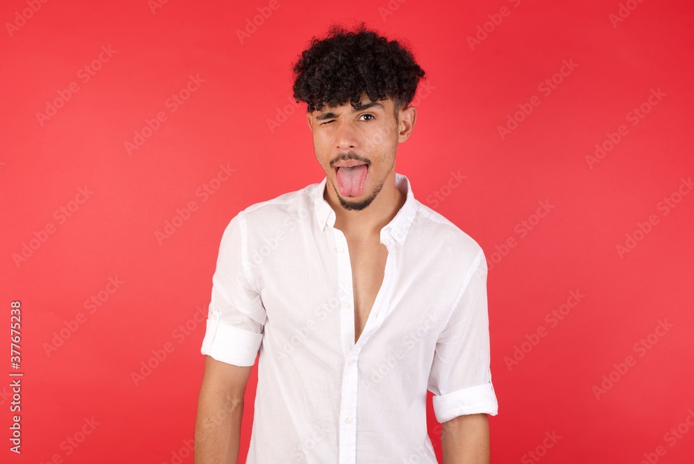 Young arab man with afro hair wearing shirt standing over isolated red background sticking tongue out happy with funny expression. Emotion concept. Young arab man with afro hair wearing shirt standing over isolated red background sticking tongue out happy with funny expression. Emotion concept.