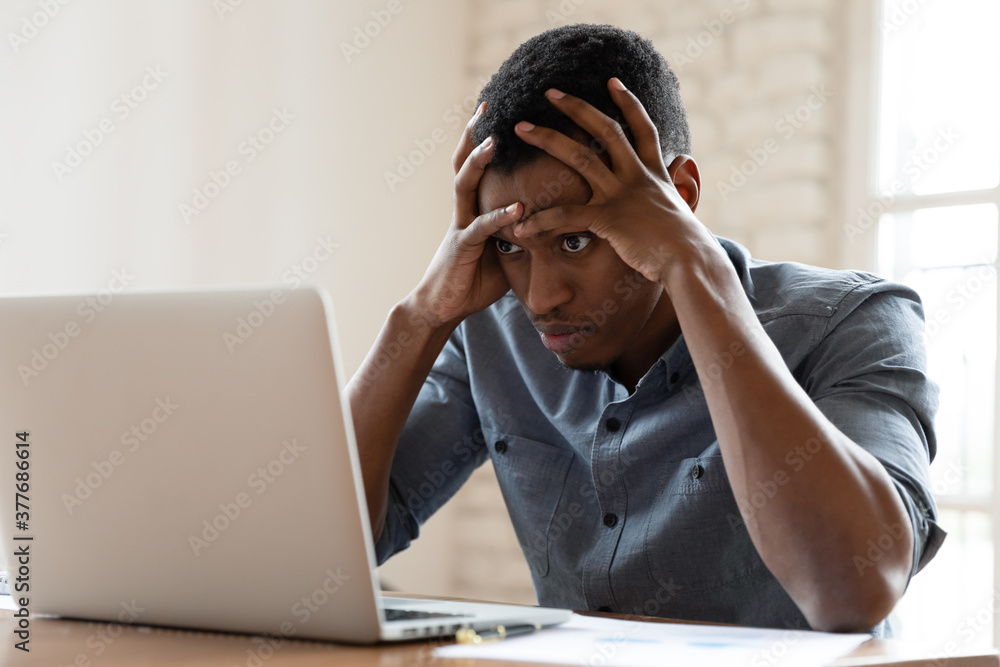 Mad African American young male employee look at laptop screen having operational software problems, angry biracial man worker work in office annoyed with slow Internet connection on computer - obrazy, fototapety, plakaty 