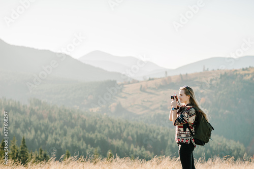 Hipster girl in sunglasses with camera and backpack travels and enjoys nature. Hiker in the mountains at sunset. Woman photographing the sunrise.