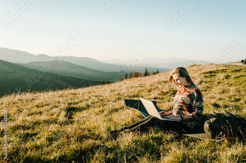 Woman traveler using laptop computer while enjoying sunset in mountains. Freelancer girl working on netbook during vacation holidays in autumn. Distant work and travel, freelance as lifestyle concept.