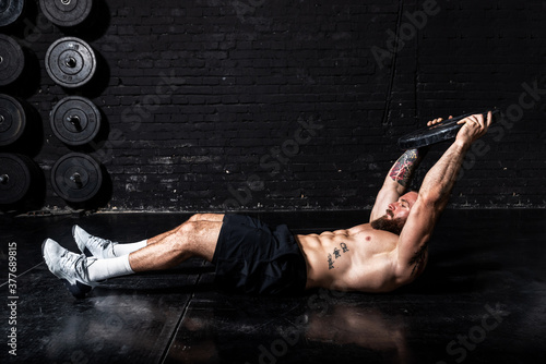 Young sweaty fit strong muscular man with big muscles doing hardcore cross abs workout training on the gym floor with weight plate