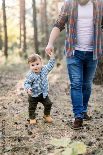 Happy fatherhood and family walk concept. Cropped image of little baby boy walking together with his young handsome father in stylish casual clothes in beautiful forest in autumn