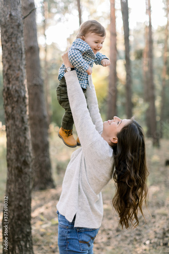 Portrait of happy Caucasian mother lifting and playing with her cute little son, enjoying their joint walk in pine forest together. Happy family concept, mom and son in forest photo