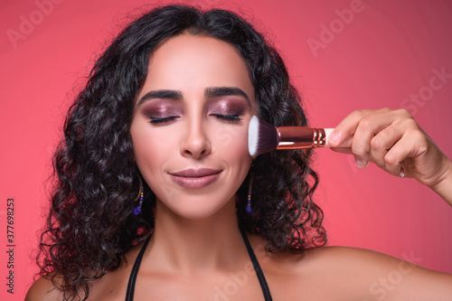 Close-up portrait of beautiful girl doing make-up. Someone is holding brush and touching it to her cheek carefully. Isolated on pink background